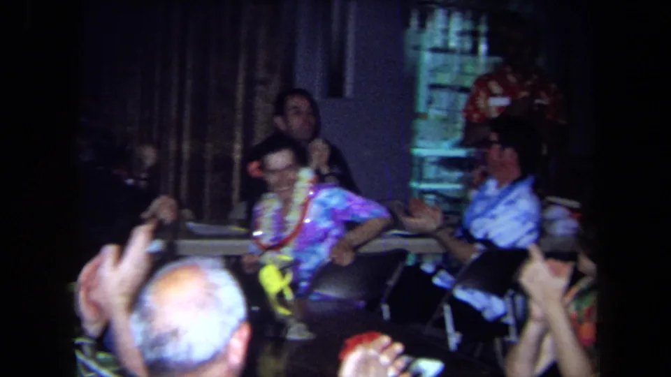 Adults wearing leis sit in folding chairs and clap during a 1971 California indoor celebration captured on 8mm film.