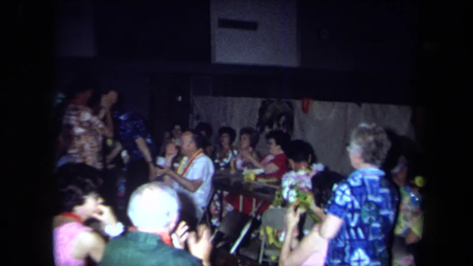 Wide 8mm view of adults seated and standing around tables and chairs at a 1971 California indoor celebration.