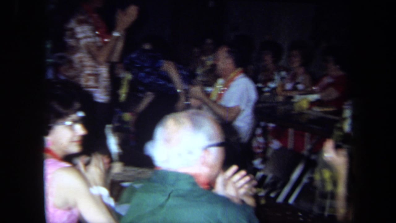1971 California home-movie frame of adults at a crowded Greek family reunion, some clapping near banquet tables in a dim interior.