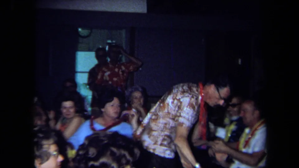 1971 California reunion scene showing adults seated in a dim hall while a man in a patterned shirt leans toward someone at a table.