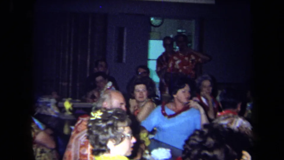 1971 California home-movie frame of adults in leis seated at reunion tables, with a standing figure near the back of the room.