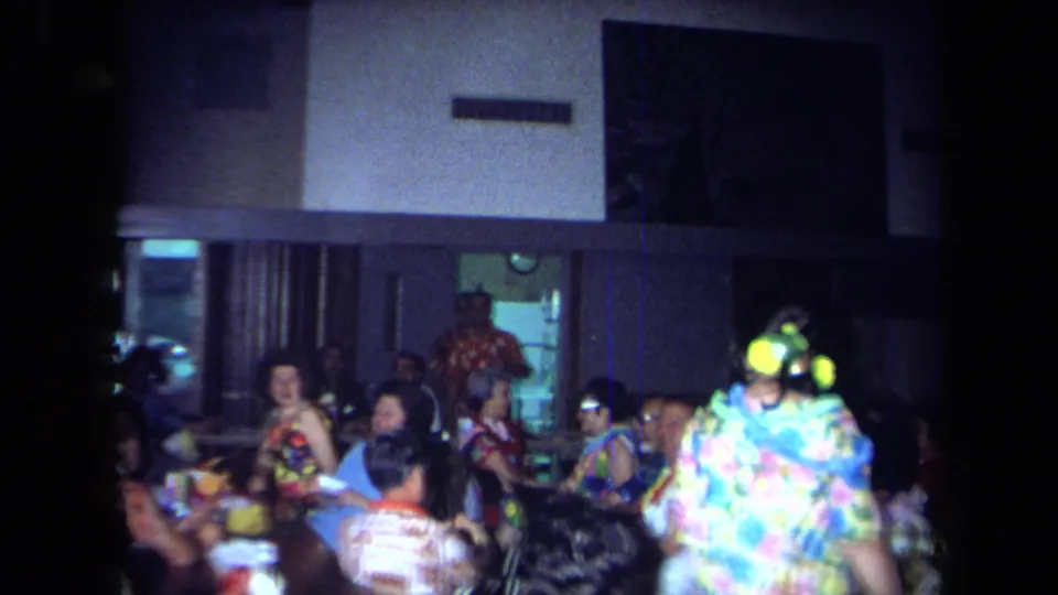 Wide 1971 California reunion frame showing seated guests at tables in a hall, with a brightly dressed person moving at the right edge.