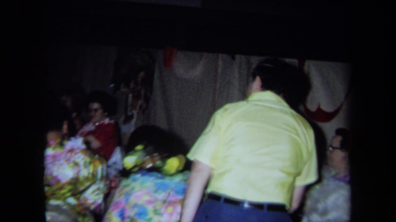 1971 California home-movie frame showing adults at a crowded indoor party with bright patterned clothing and a man standing near seated guests.