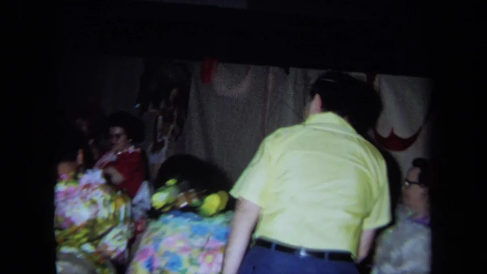 1971 California home-movie frame showing adults at a crowded indoor party with bright patterned clothing and a man standing near seated guests.