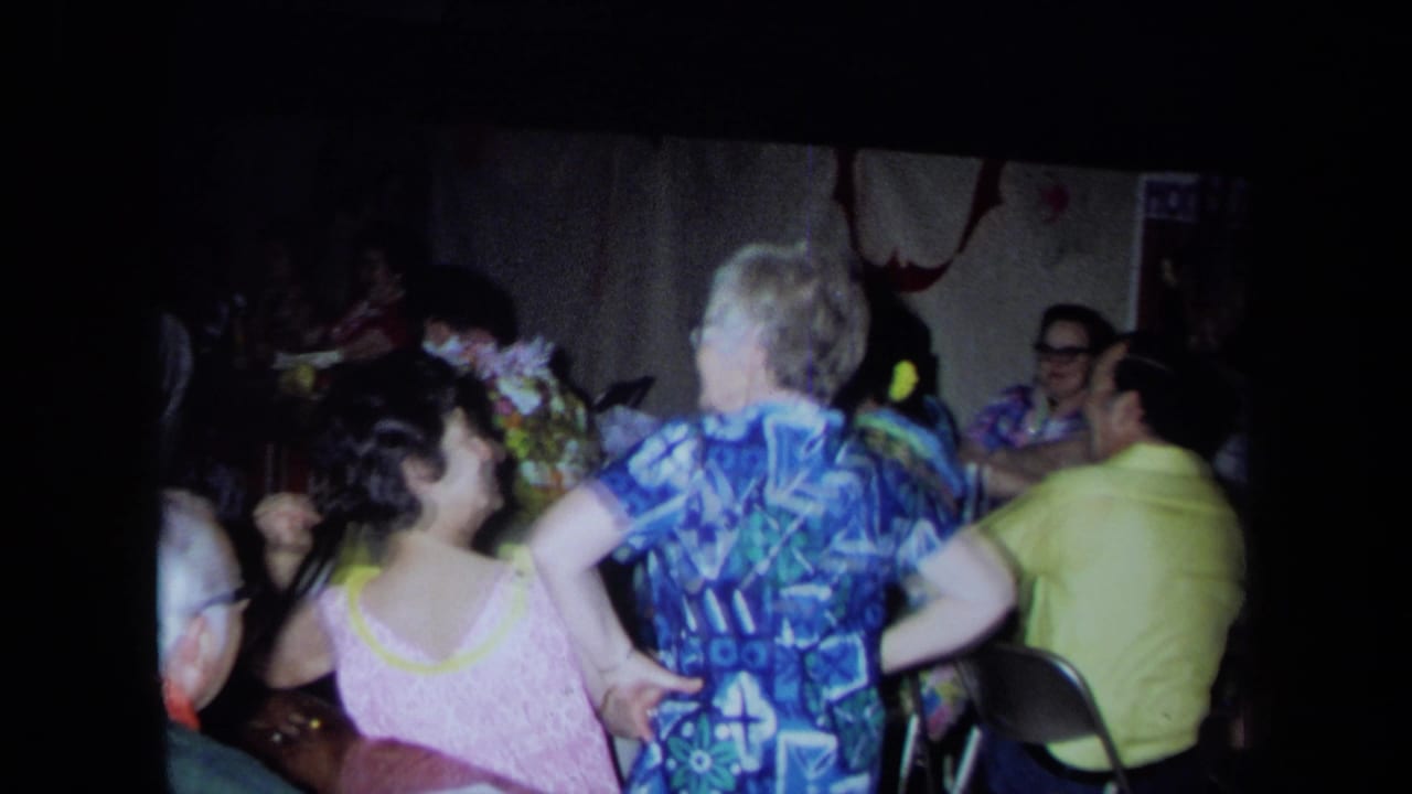 Two women dance or stand near seated guests in a crowded indoor party room.