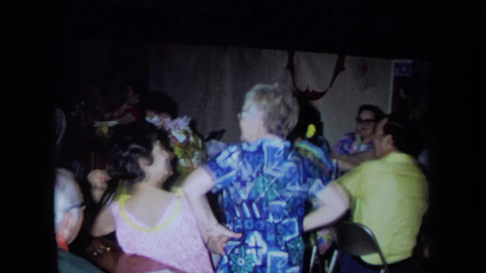 1971 California home-movie frame with a woman in a blue patterned dress and another in pink near seated party guests.