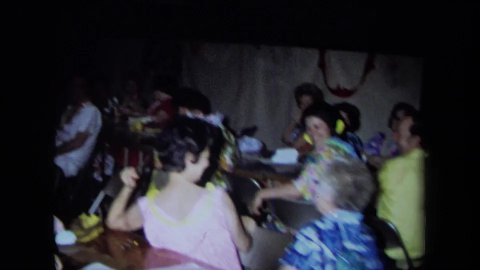 1971 California home-movie frame showing a crowded private party with seated adults, tables, and women moving in the foreground.