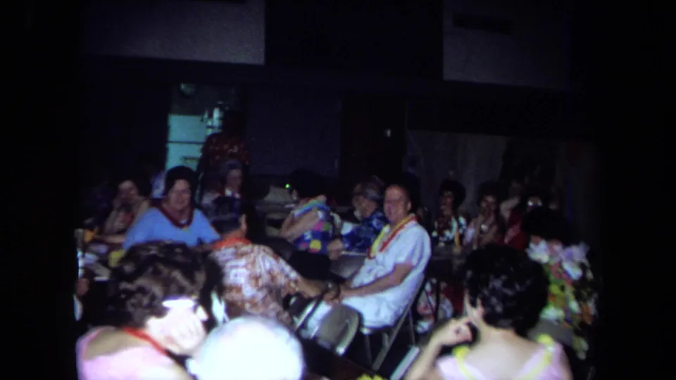 1971 California home-movie frame showing seated adults in a dim private party interior with colorful clothing and tables.