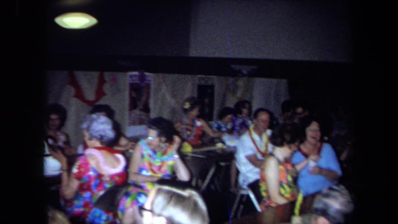 1971 California home-movie frame of adults at an indoor party, seated around tables with leis, patterned clothing, and dim overhead light.