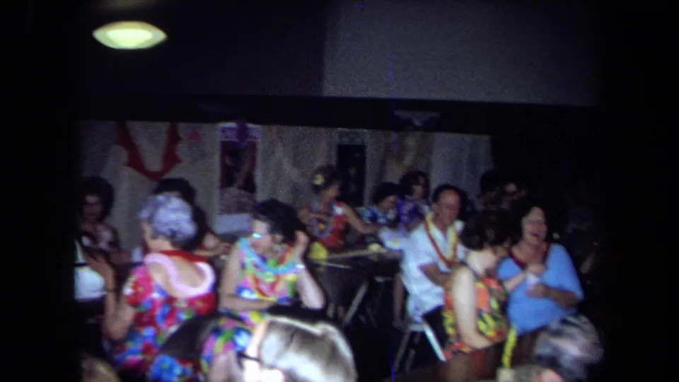 1971 California home-movie frame of adults at an indoor party, seated around tables with leis, patterned clothing, and dim overhead light.