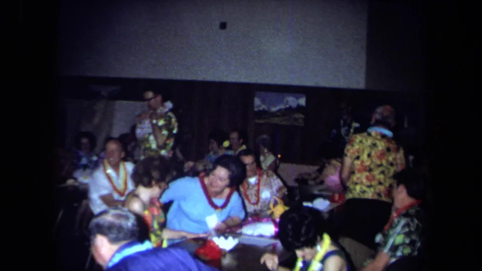 Adults in leis and bright shirts gather in a California indoor party scene, with tables, wall art, and handheld 8mm framing.