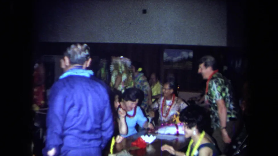 Home-movie view of adults at an indoor 1971 California celebration, with a man in a blue sweater and guests in leis near tables.