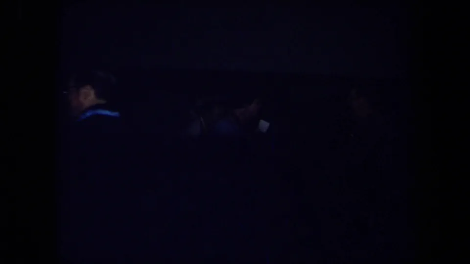 Dark 8mm frame from the indoor party, with only faint silhouettes of guests and the room visible due to low exposure.