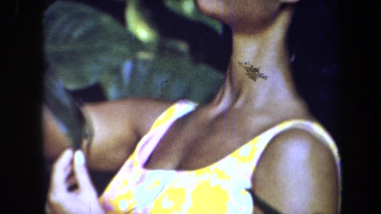 1971 8mm frame from California showing a posed woman in a yellow floral dress partly hidden by large green leaves.