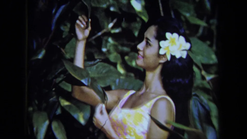 1971 California 8mm frame showing a woman with a white plumeria-like flower in her hair reaching into leafy greenery.