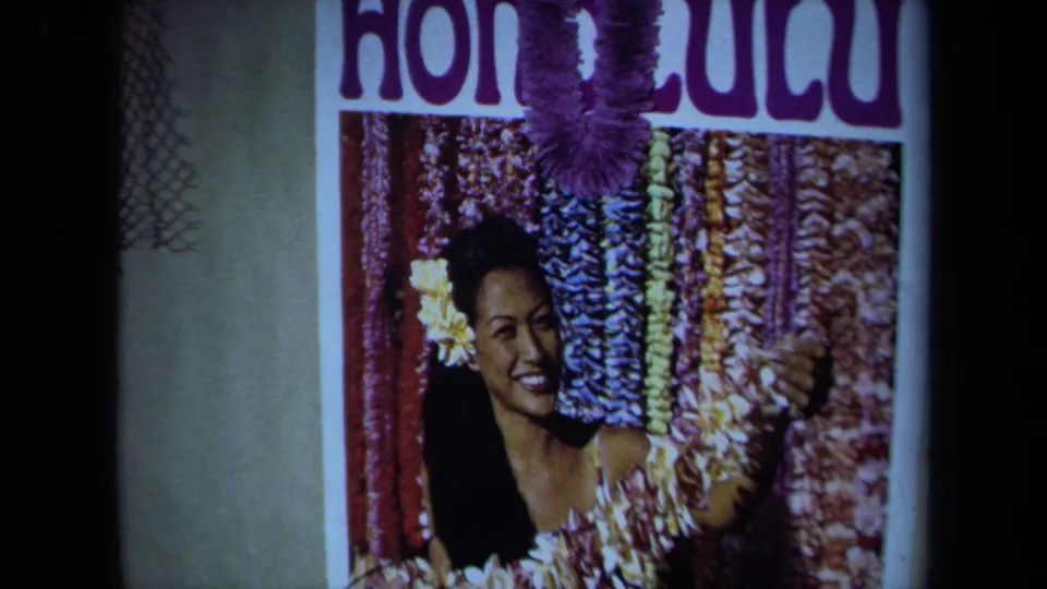 1971 California 8mm frame of a Honolulu-themed poster with a smiling woman, flower adornment, and hanging leis.