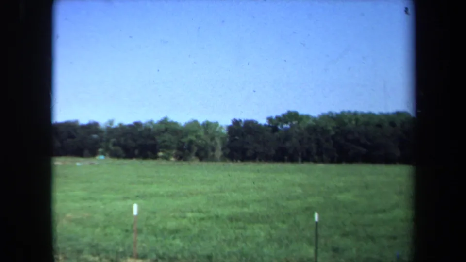 1971 California 8mm frame of an open green field with small posts, a distant tree line, and clear blue sky.