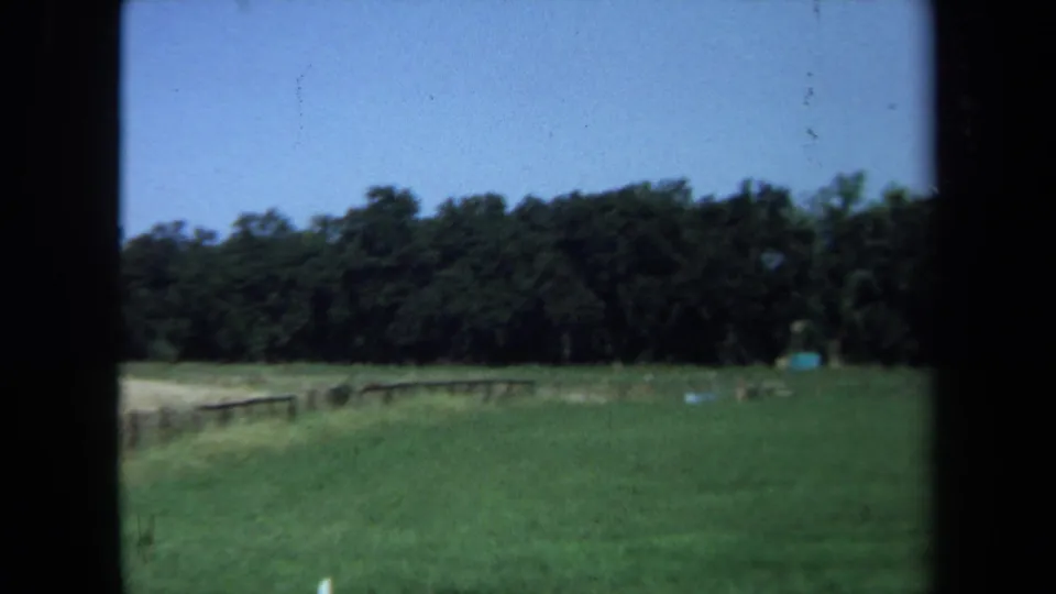 1971 California 8mm frame with a green field, low wooden fence, distant trees, blue sky, and soft home-movie focus.