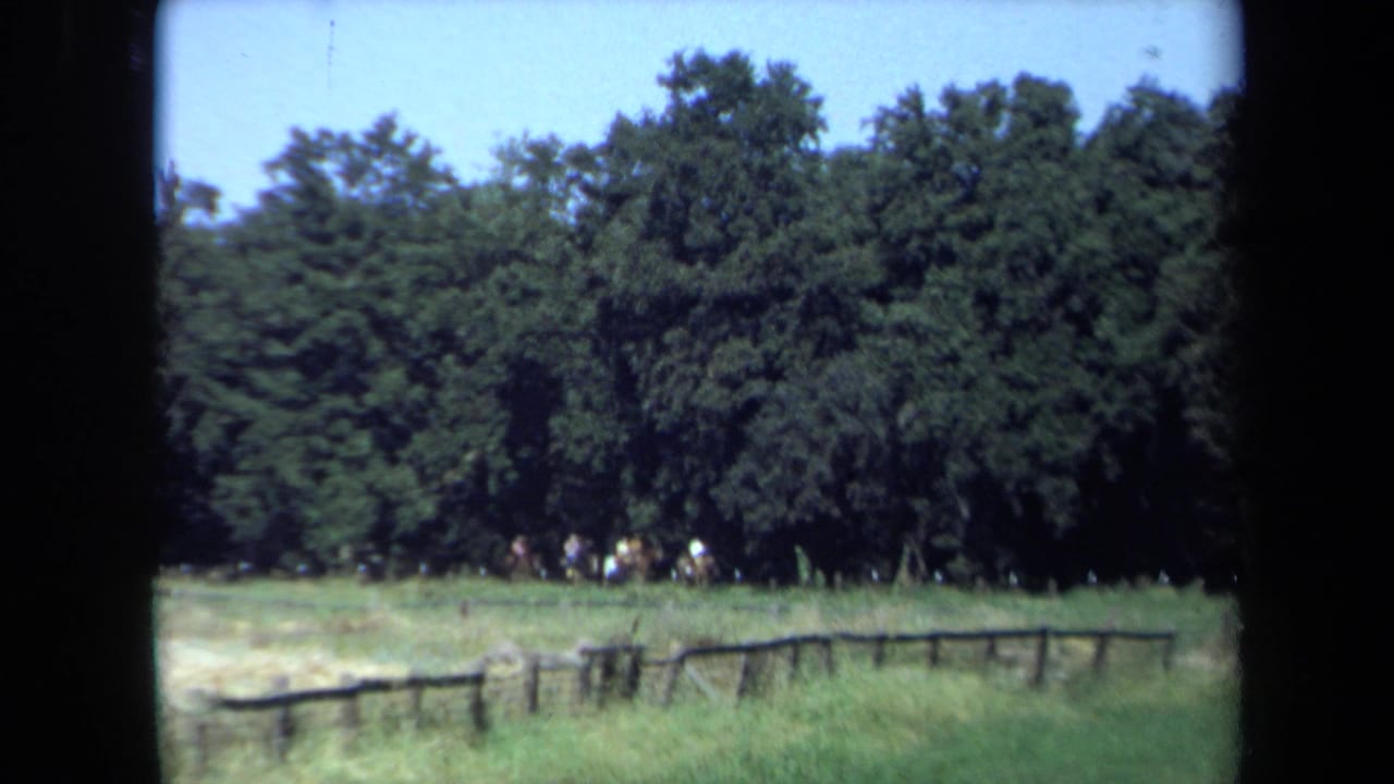 1971 California 8mm frame showing distant horseback riders at the edge of a green clearing, with trees, blue sky, and a low wooden fence.