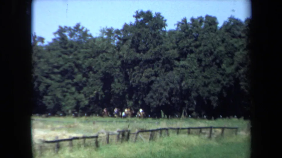 1971 California 8mm frame showing distant horseback riders at the edge of a green clearing, with trees, blue sky, and a low wooden fence.