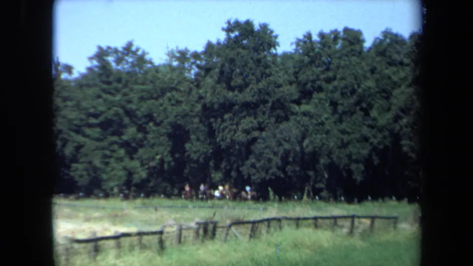 Distant riders and horses appear near dense trees in a California clearing, framed by 8mm film edges and a foreground fence.