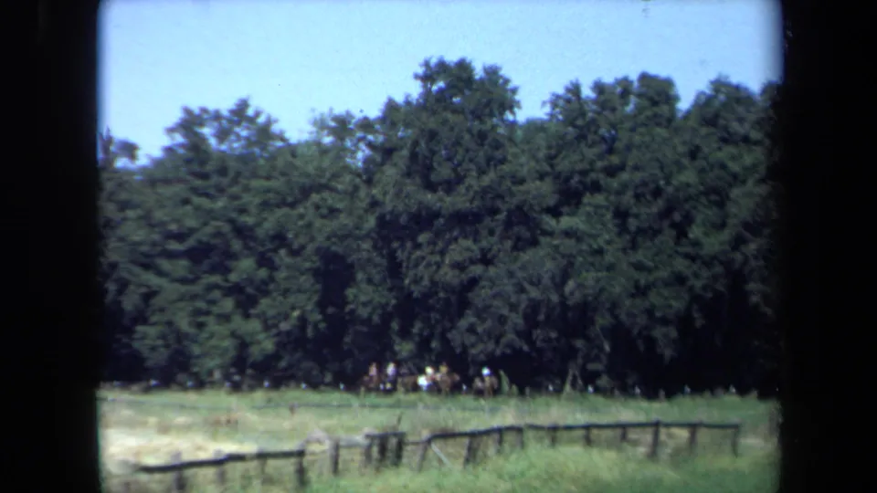 1971 home-movie frame of riders clustered along a wooded edge, with a weathered fence crossing the foreground of the California field.