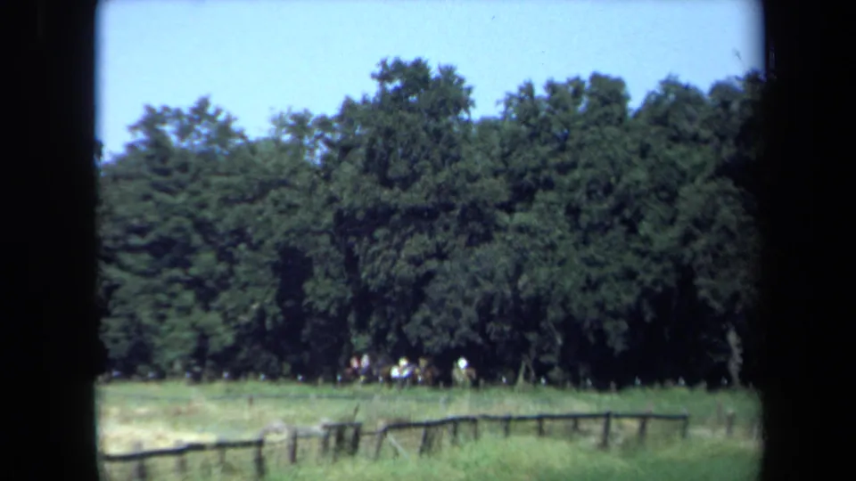 Distant horseback riders remain near the wooded edge of a green California clearing, with a low fence and saturated 8mm color visible.