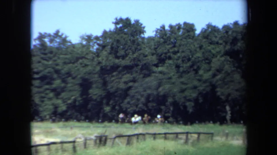 Vintage 8mm view of riders near a dense California tree line, seen from across a fenced grassy field.