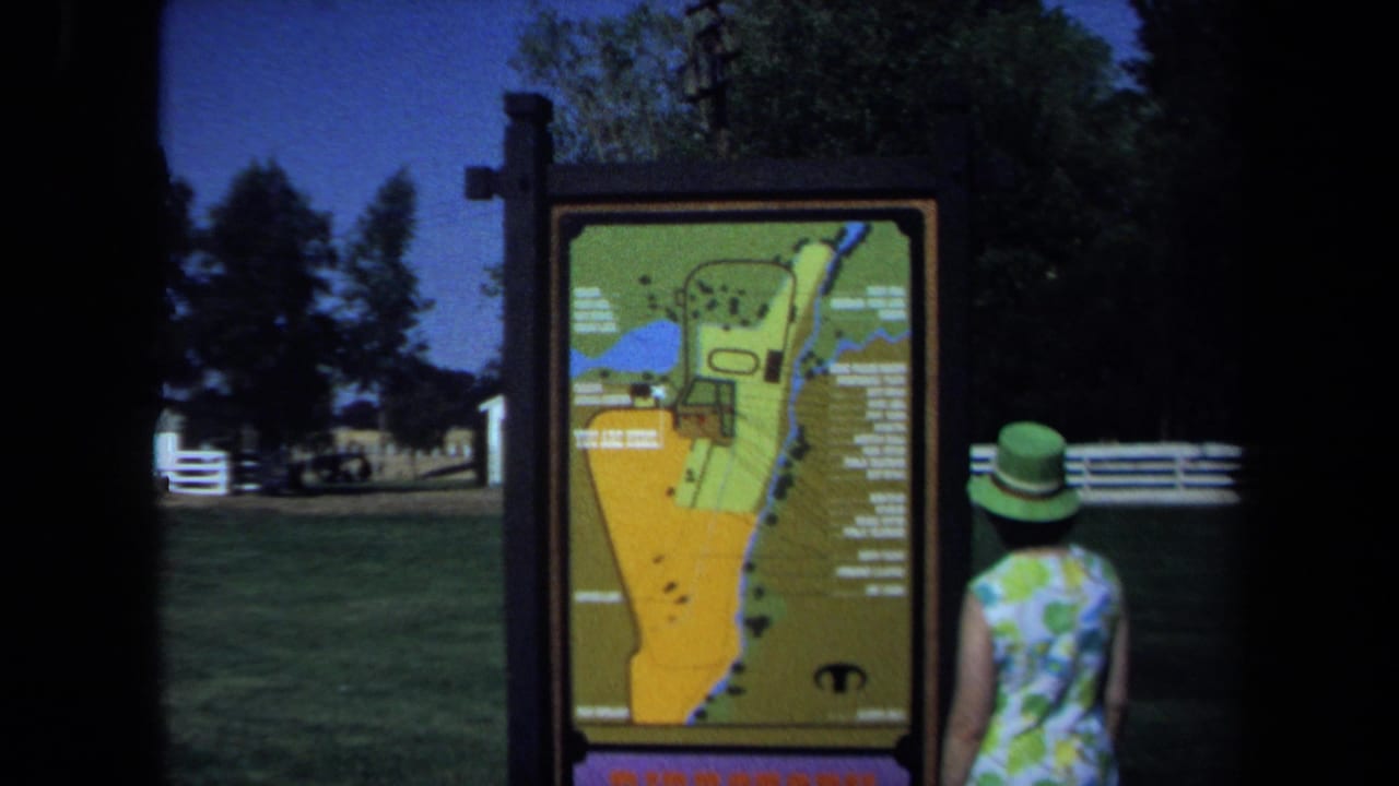 In 1971 California park footage, a woman in a green hat and floral dress looks toward a large illustrated map sign near grass and trees.