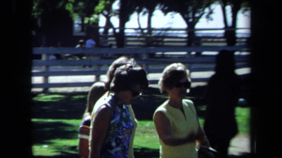 1971 California park scene with two women in sunglasses walking together on a sunny lawn, with trees and rail fencing behind them.