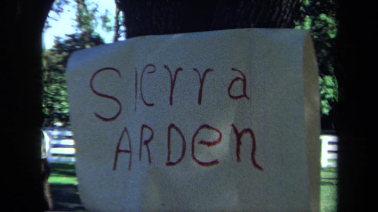 1971 California 8mm frame showing a paper sign reading Sierra Arden against dark tree trunks with a white fence beyond.