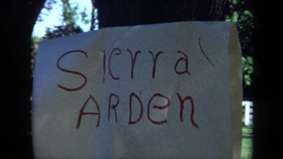The Sierra Arden paper sign remains centered on the tree, with bright yard foliage and a white fence behind it.