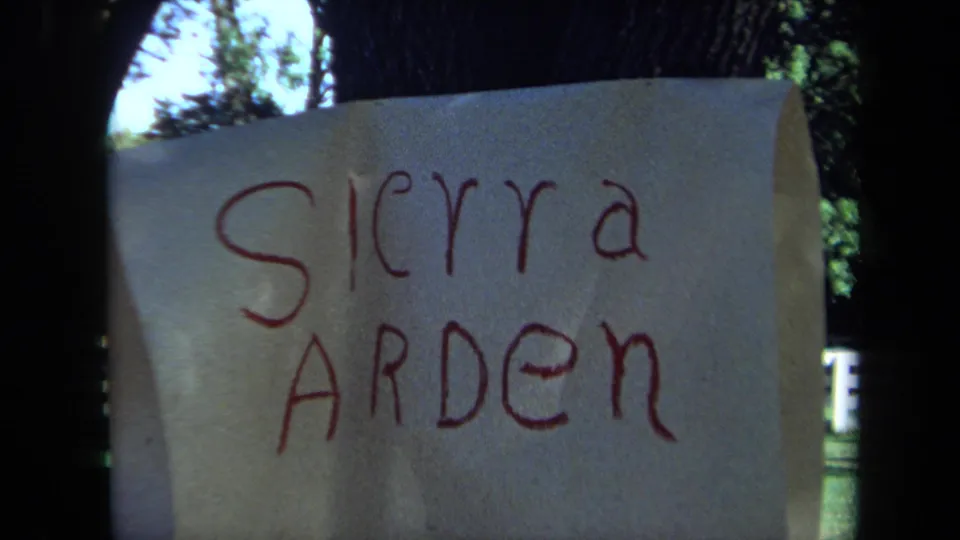 The paper sign reading Sierra Arden fills the frame, with slight curl at the edge and a fenced yard visible behind.