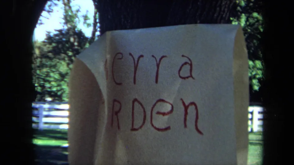 The paper has curled over part of the Sierra Arden lettering, revealing the yard, tree trunks, and white fence beyond.