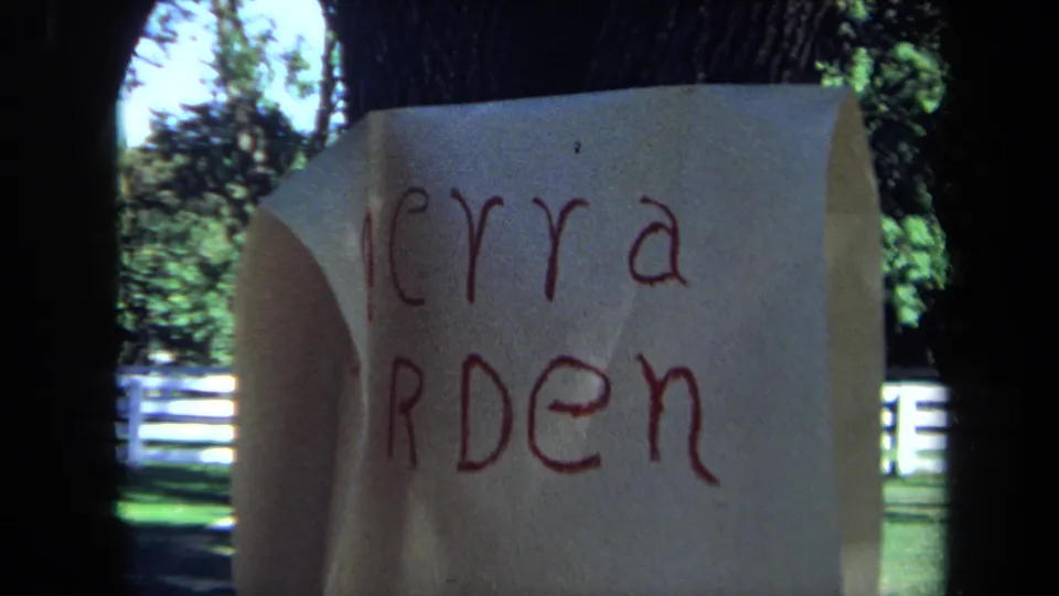 1971 California 8mm frame with a handwritten Merry Garden note in the foreground and park trees and white fencing behind it.