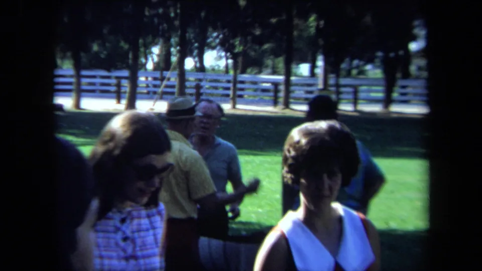 1971 California home-movie frame showing adults crossing a shaded lawn in a park with trees and white fencing in the background.