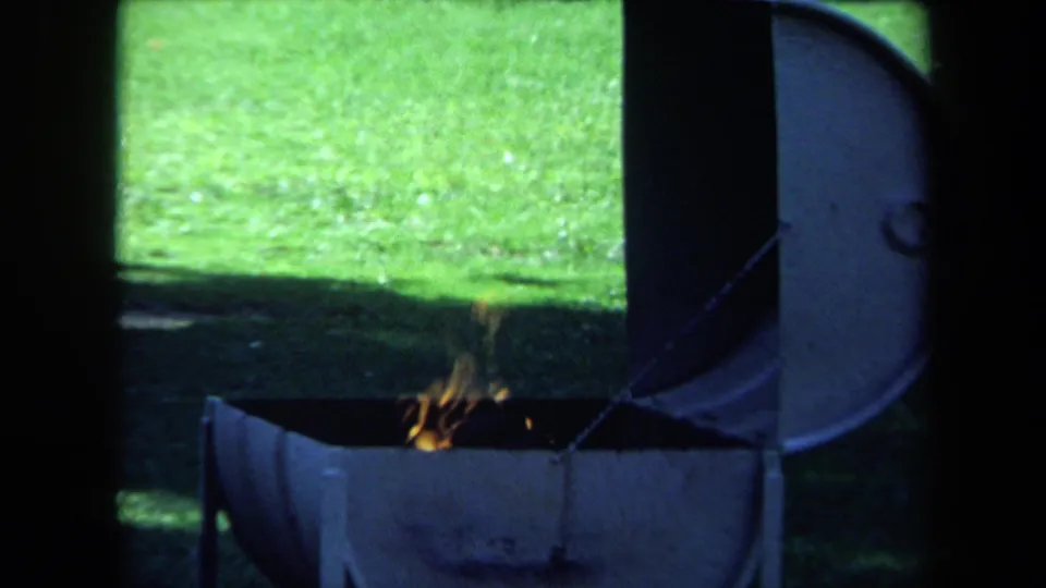 1971 California 8mm frame showing a small fire in an open metal barrel beside green grass and tree shade.
