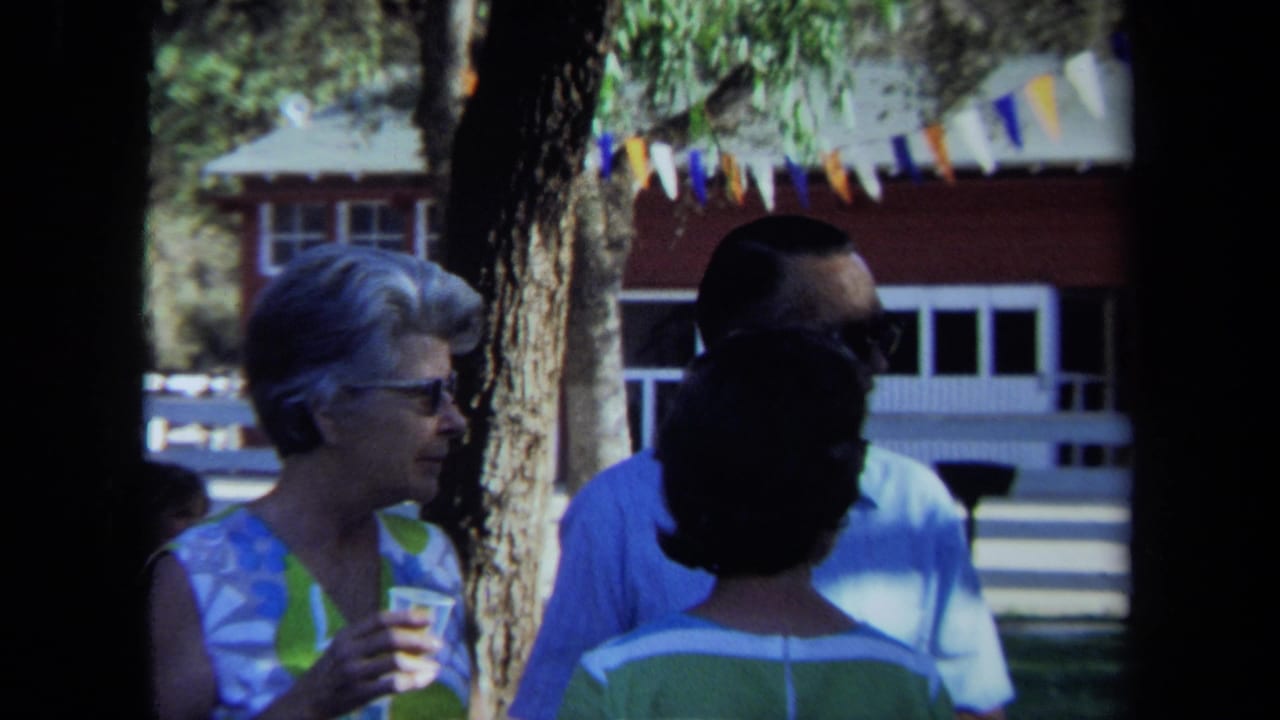 Three adults talk outdoors near trees, with a red building and pennant flags behind them.