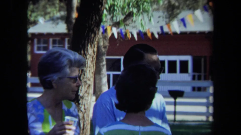 1971 California frame showing adults in conversation under trees, with colorful pennants, a white fence, and a red building behind them.