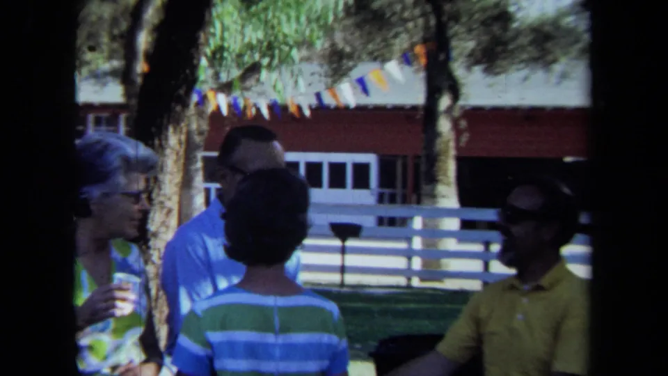 1971 California home-movie frame with four adults gathered outdoors, including a man in a yellow shirt joining the conversation.