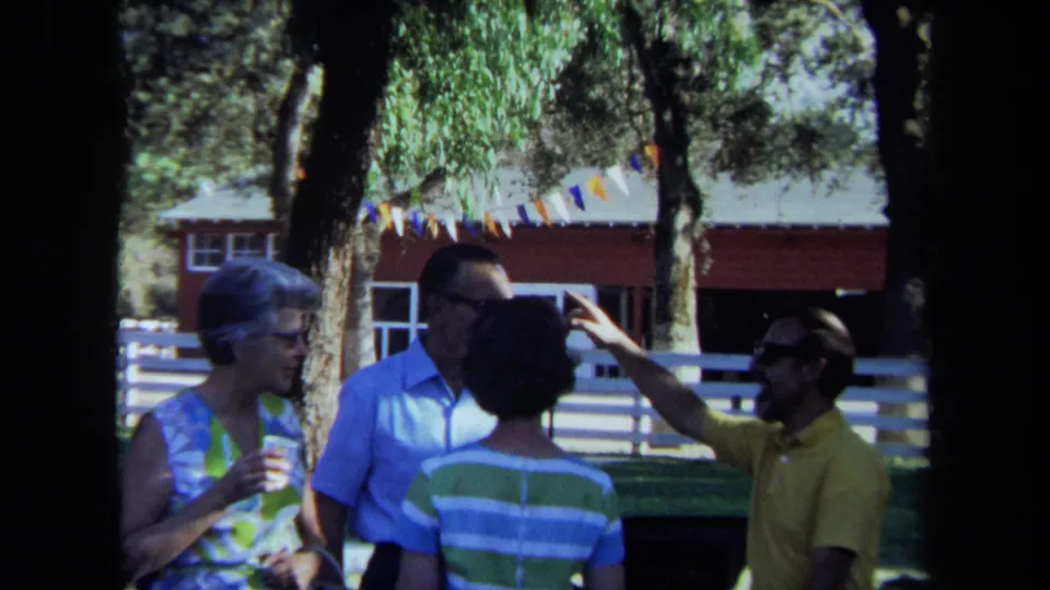 1971 California outdoor gathering frame with a man gesturing as adults converse beside trees, pennants, a white fence, and a red building.