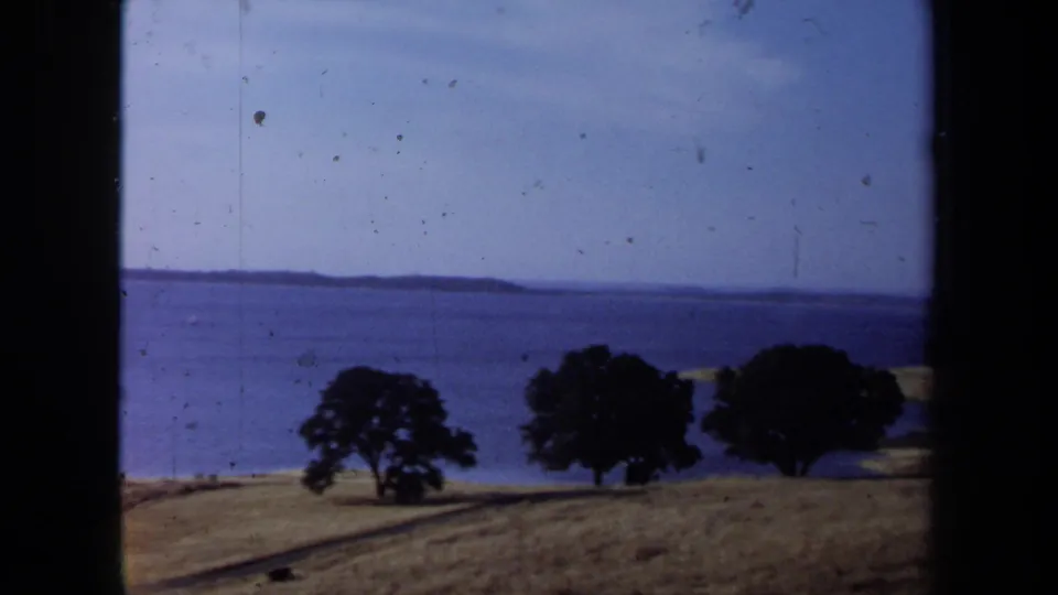 1971 California coastal view with dry grass, dark trees, blue ocean, and distant land across the water.