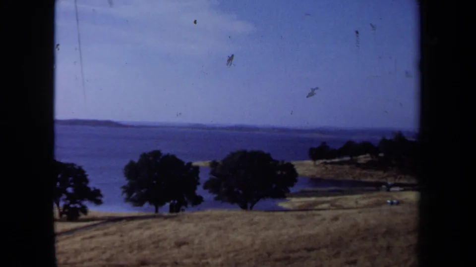1971 California 8mm view of a grassy slope, dark trees, shoreline, and blue water under clear sky.