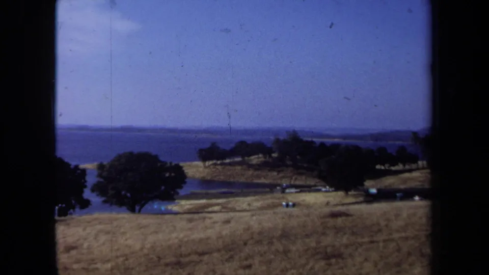 1971 California home-movie frame showing a dry coastal hillside, trees, blue water, distant shoreline, and small cars.