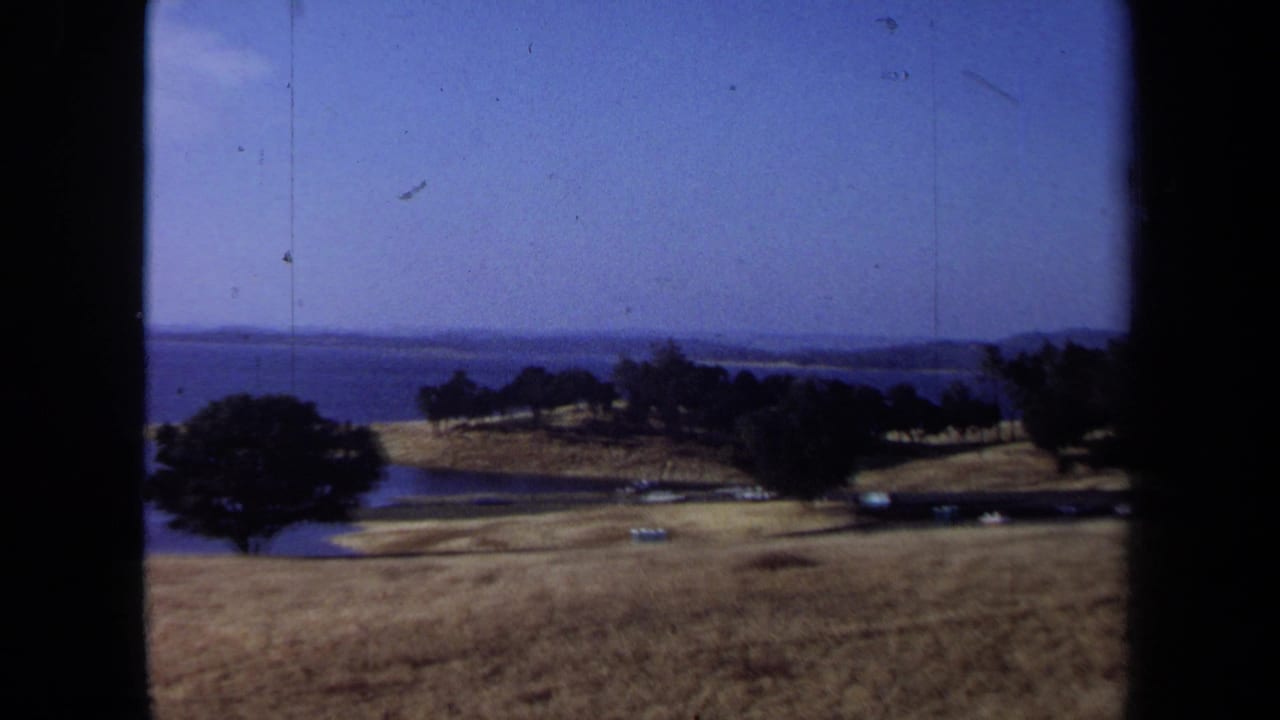 1971 California 8mm frame showing a dry grass shoreline, trees, calm blue water, and distant land under a pale sky.