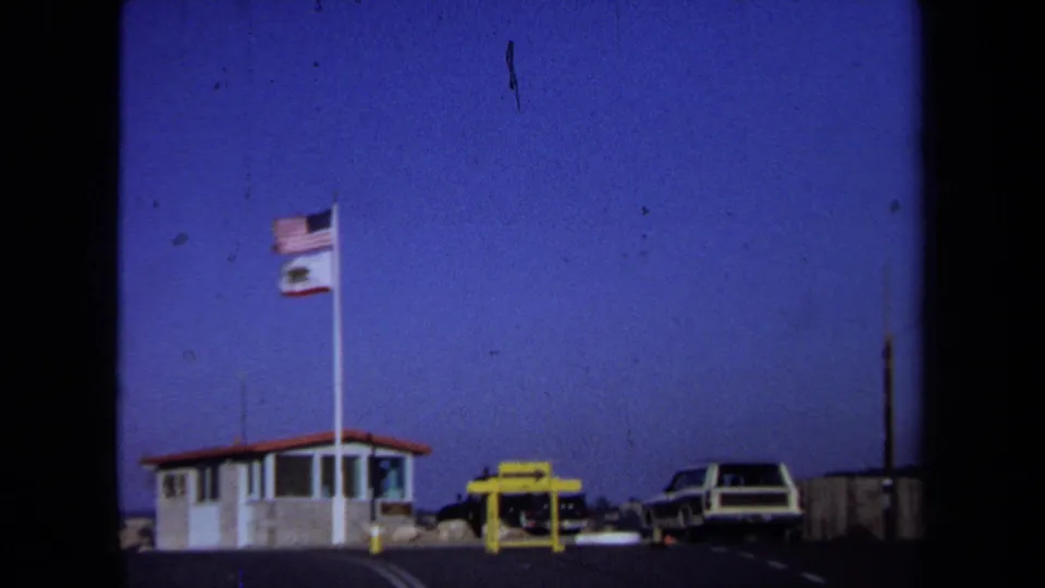 1971 California roadside rest-stop view with a small booth, tall flagpole, yellow sign, and parked 1970s vehicles.