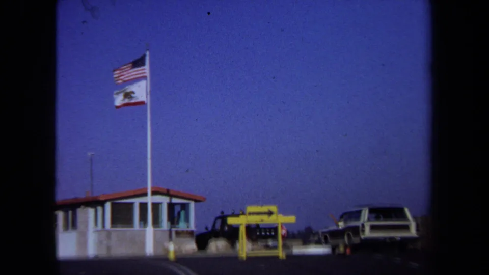 California rest-stop frame from 1971 showing state and national flags, a low booth, yellow sign, and a station wagon.