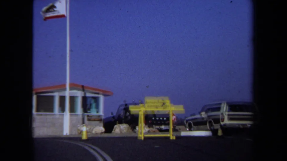 1971 California 8mm view of the rest-stop booth, flags, yellow sign, roadside rocks, and parked vehicles.