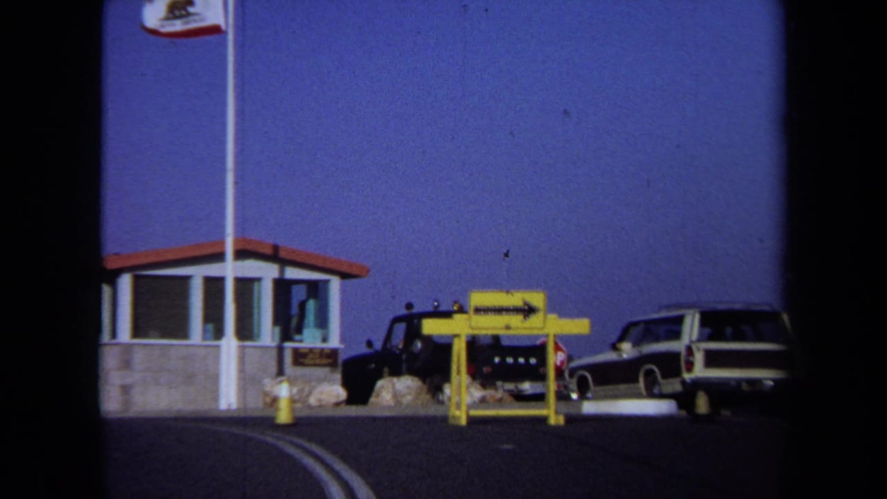 1971 California 8mm frame showing a roadside entrance booth, California flag, parked cars, and a yellow sign near the curb.