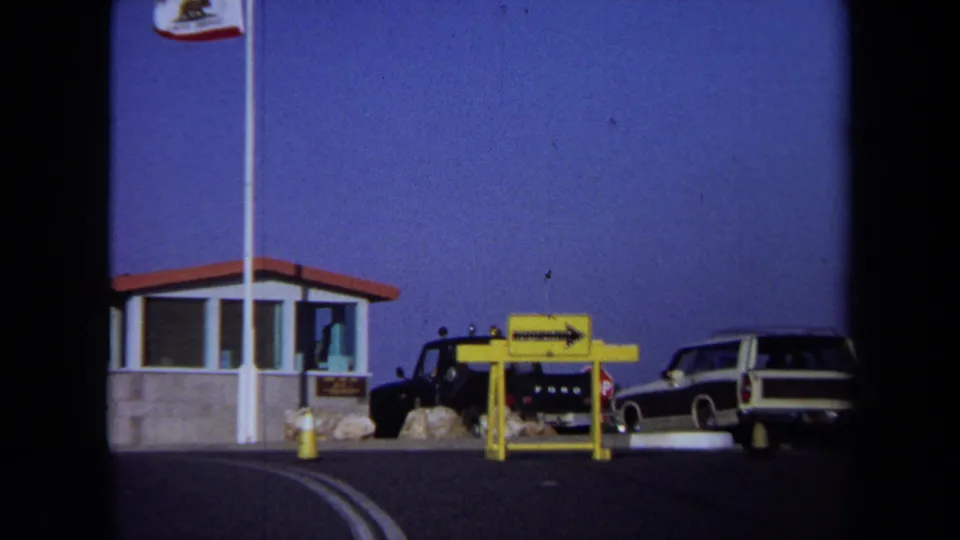 1971 California 8mm frame showing a roadside entrance booth, California flag, parked cars, and a yellow sign near the curb.